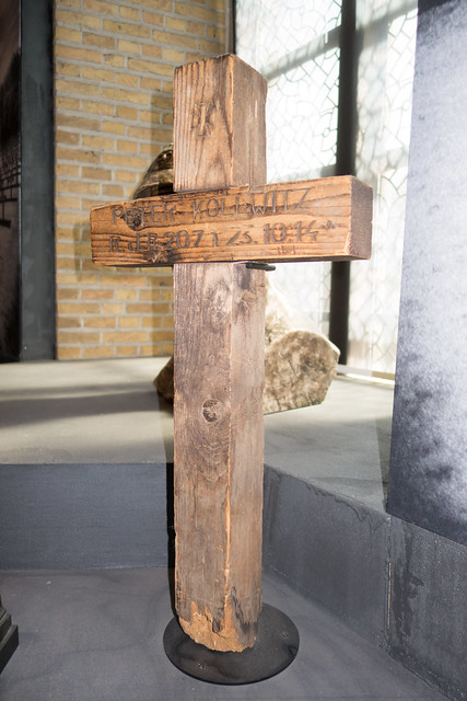 A weathered, wooden cross stands on a black base inside a room with brick walls and large windows. Carved text is visible, conveying an aged, solemn tone. Peter Kollwitz grave marker.
