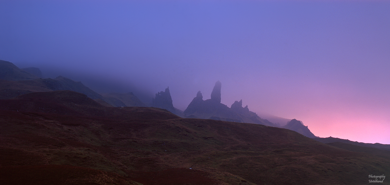 Misty mountains with jagged peaks silhouetted at dawn, under a purple sky transitioning to pink. The scene evokes a serene, mystical atmosphere. The Ols Man of Storr, Trotternish Ridge, Isle of Skye.