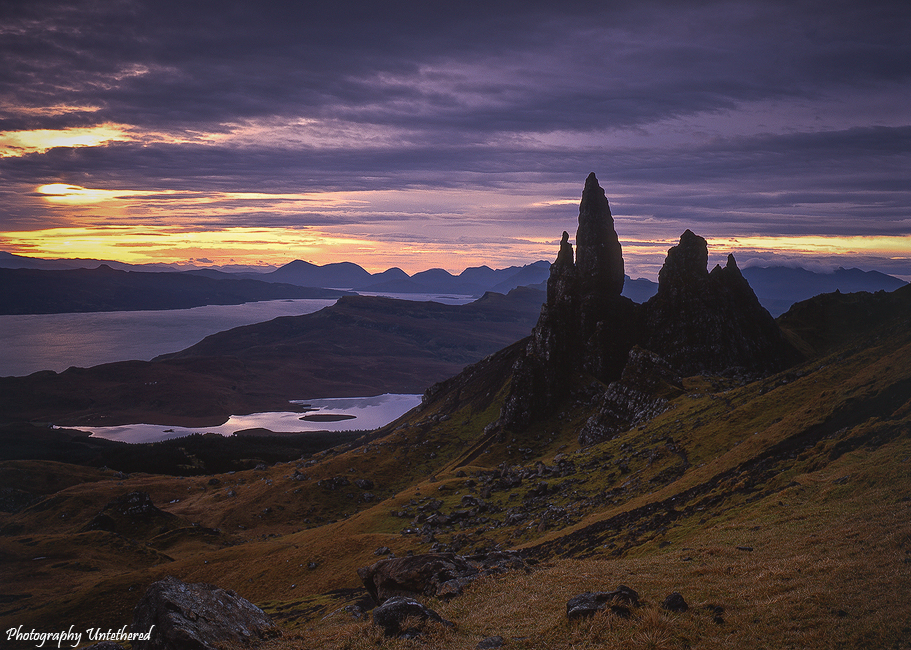 Dramatic landscape at sunset with jagged rock formations in the foreground, vibrant orange sky, and a calm sea surrounded by distant mountains. Old Man of Storr, Trotternish Ridge, Skye