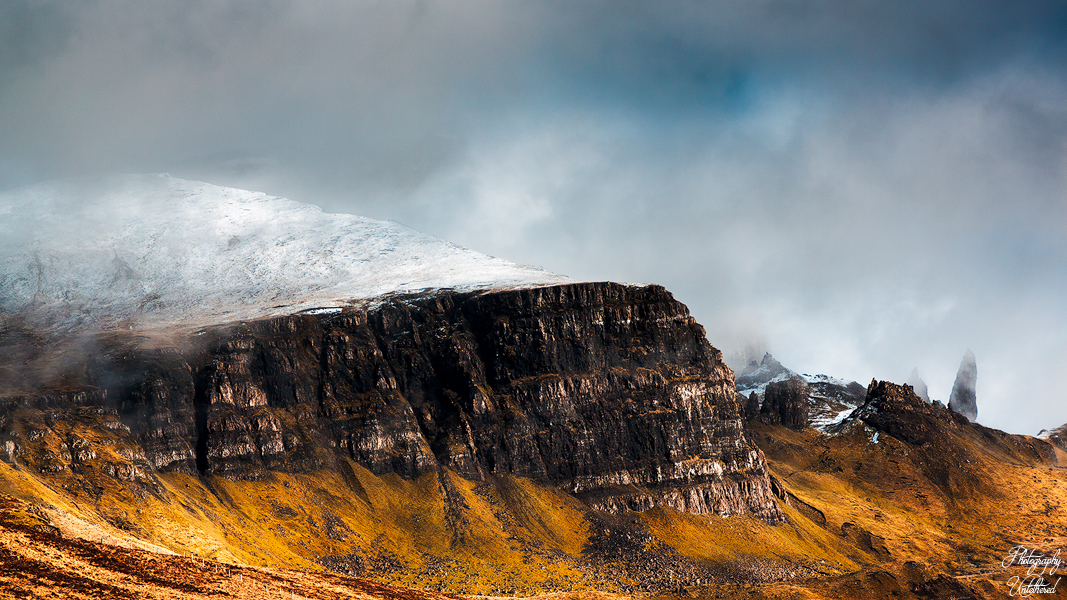 Dramatic landscape of a rugged mountain with a snow-dusted peak, steep rock faces, and golden grassy slopes under a moody, overcast sky. The Storr (Scottish Gaelic: An Stòr) is a mountain on the Trotternish peninsula of the Isle of Skye in Scotland.