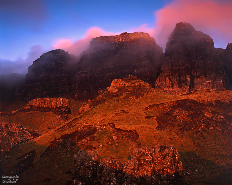 Majestic mountains with rugged cliffs shrouded in soft pink clouds at dusk, warm sunlight casting a golden glow on the rocky landscape below. Sunrise The Storr, Skye.