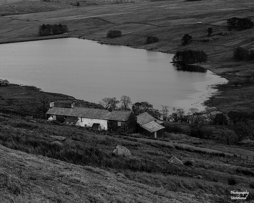 Black and white rural landscape showing a serene lakeside view with a rustic stone cottage nestled in rolling hills. The scene feels calm and timeless. Uncle Monty’s Crow Crag Seddale Hall. Photography Untethered.