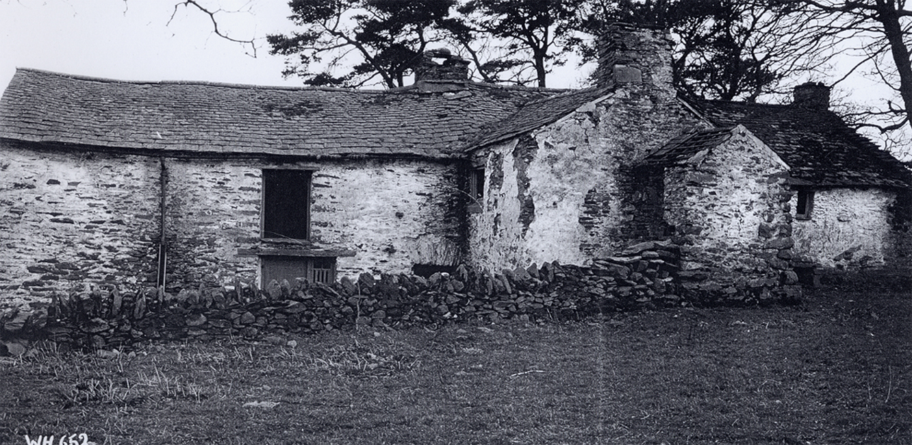 Old stone cottage with a weathered, uneven roof, partially crumbling walls, and surrounded by a stone fence. Leafless trees rise in the background.