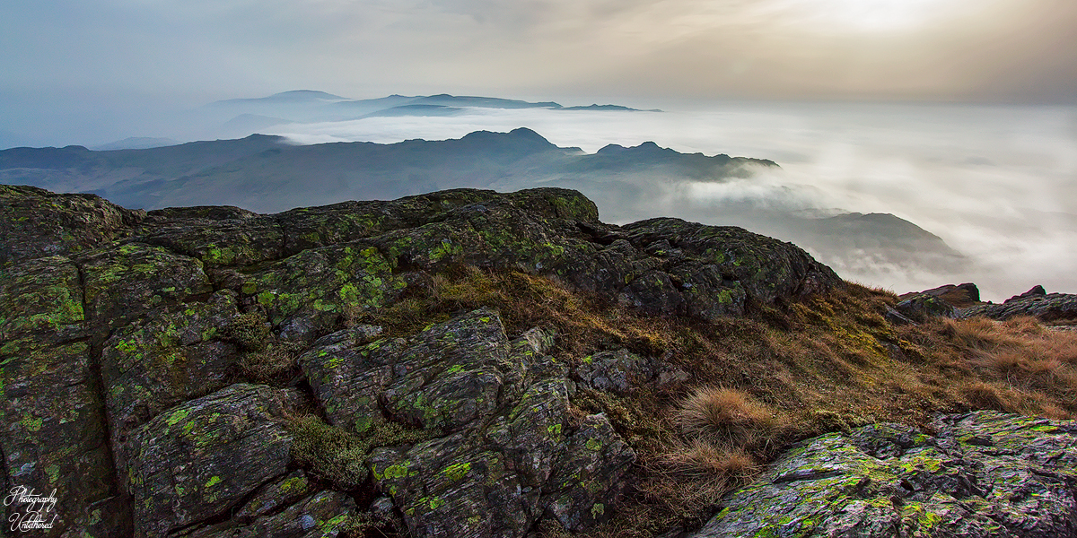 Rocky terrain covered with moss in the foreground, overlooking a mist-filled valley and distant hills under a soft, early evening cloudy sky.