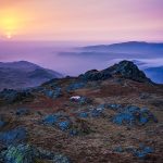 A serene sunrise over a rugged mountain landscape, with a vibrant purple and orange sky. Soft mist lingers between distant hills, creating a tranquil scene. Harter Fell, Eskdale.