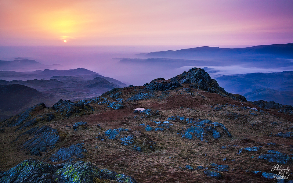 A serene sunrise over a rugged mountain landscape, with a vibrant purple and orange sky. Soft mist lingers between distant hills, creating a tranquil scene. Harter Fell, Eskdale.