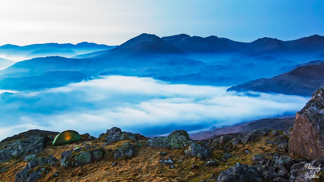 A green tent sits on rocky terrain above a blanket of clouds, with majestic mountains in the distance under a serene blue sky, evoking tranquillity and adventure. Harter Fell, Eskdale.