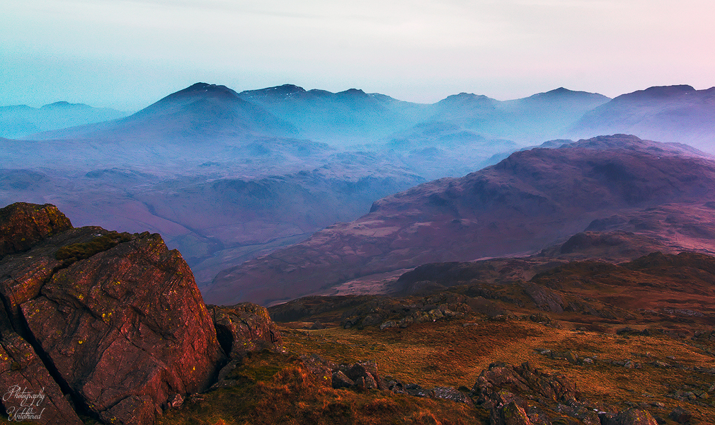 Serene mountain landscape at dusk, with rocky foreground bathed in warm light. Misty blue peaks recede into the distance under a soft pink sky.