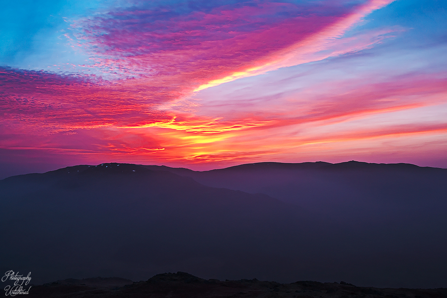 Vivid sunset over dark mountains, featuring a sky painted with bright orange, pink, and blue hues. The scene conveys a serene and majestic atmosphere—Sunrise over Grey Friar and the Coniston Hills from Harter Fell.