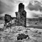A moody black-and-white photo of ancient stone ruins in a desolate landscape. Dark clouds loom overhead, evoking a sense of mystery and timelessness. Ardvreck Castle, Assynt, Scotland.