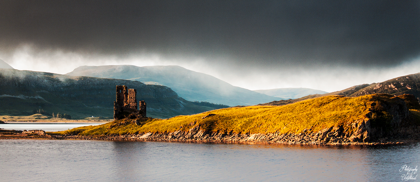 A dramatic landscape with a ruined Ardvreck Castle on a sunlit hill beside a dark, cloudy sky. The scene is framed by serene water and distant mountains.
