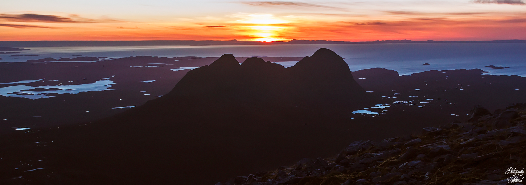 Sunset over Suilven Mountain silhouette against a vibrant sky, with orange and purple hues. Calm lochs, sea and rugged terrain create a serene, majestic atmosphere.