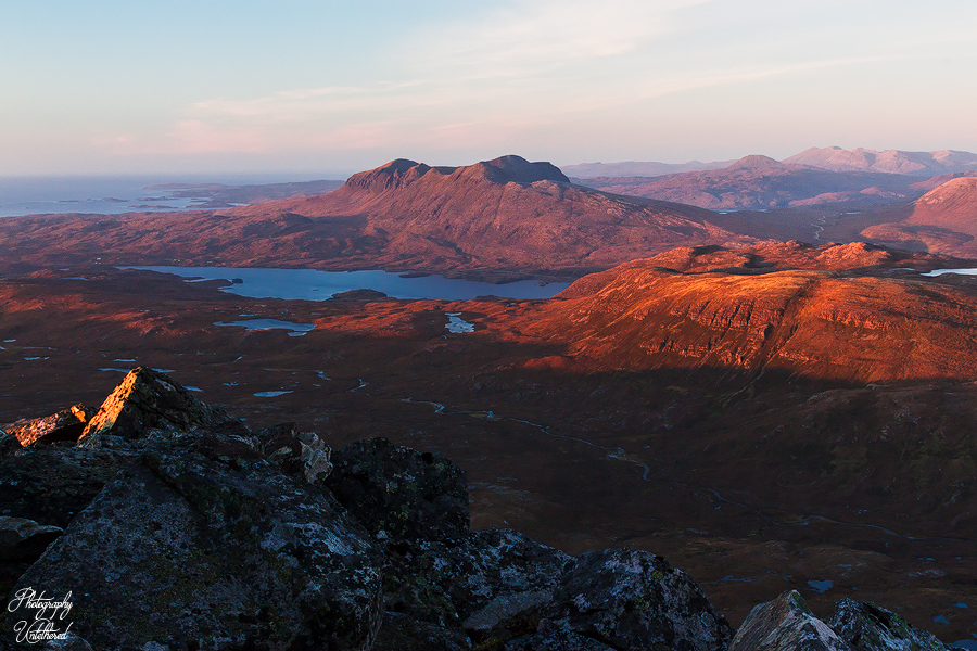 Overlooking a mountainous landscape at sunset, with warm orange and red hues illuminating the rugged terrain and a serene lake below. Peaceful ambience. Overlooking a mountainous landscape at sunset, with warm orange and red hues illuminating the rugged terrain and a serene lake below. Peaceful ambience. View of Quinag from Canisp.