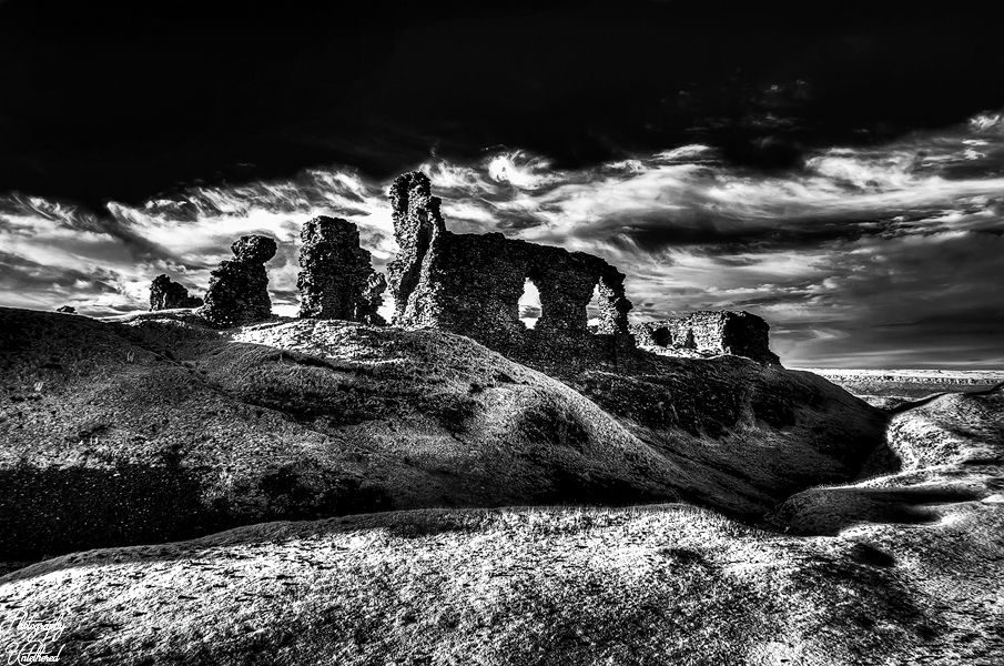A dramatic black and white image of the ancient stone ruins of Castell Dinas Brân under a cloudy sky. The scene conveys a sense of mystery and timelessness. Black and White Photography.