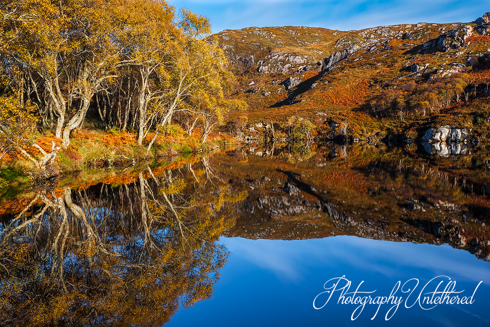 Calm lochan reflects autumn trees and rocky hills under a clear blue sky. Vibrant colors create a serene, picturesque landscape. Photography Untethered.