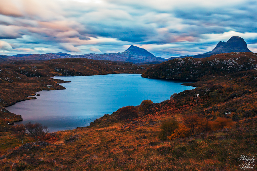 Mountainous landscape at dawn with a serene lake reflecting the vibrant sky. Rugged terrain and foliage in autumn hues, evoking tranquillity and awe. Stormy Sky over Loch Bad na Goibhre, Canisp & Suilven.