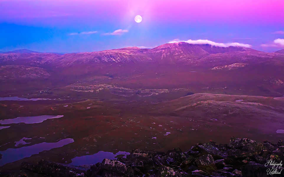 A full moon illuminates a serene mountainous landscape at dusk, casting a violet and pink hue over the terrain. Small lakes dot the scene, evoking a tranquil, mystical atmosphere. Twilight: Conival and Ben More Assynt from Canisp