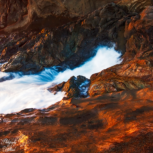 Waves crash against dark, jagged rocks, illuminated by warm, golden sunlight. The scene conveys a dynamic and dramatic seaside atmosphere. Ajuy.