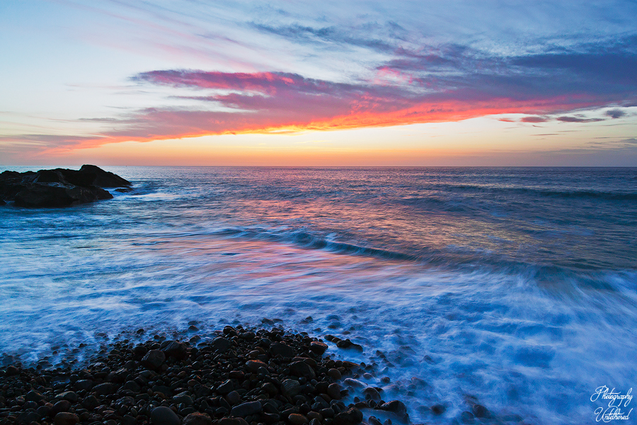 Sunset over a rocky beach with waves gently lapping the shore. The sky is painted in vibrant hues of orange, pink, and blue, creating a serene atmosphere. Ajuy Fuerteventura.