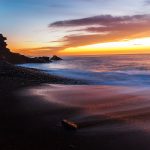 Sunset beach with dark sand, gentle ocean waves, and vivid orange and yellow sky. Rocky cliff silhouette on left; peaceful and serene atmosphere. Ajuy Fuerteventura.