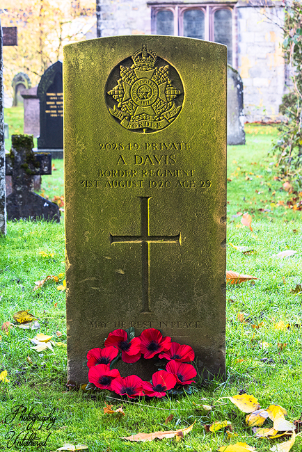 A stone war memorial with engraved names is surrounded by wreaths of red poppies. The setting conveys remembrance and honour in a solemn tone. Gravestone of Anthony Davis Border Regt; St Michael's, Shap.