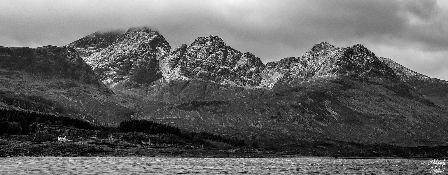 Black and white image of rugged mountains under cloudy skies. A small white house contrasts with the dark landscape, creating a dramatic, moody scene. Bla Bheinn and Loch Slapin, from Torrin | Isle of Skye. Black and White Photography.