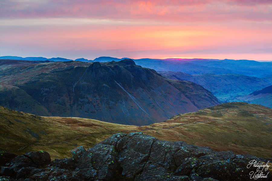 Sunrise over a vast mountain landscape, (Pike of Stickle) with rugged peaks and rolling hills. The sky glows with shades of pink and orange, creating a serene atmosphere.