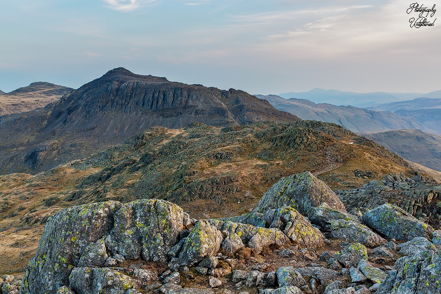 Rocky mountain landscape with rugged peaks under a soft, cloudy sky. Foreground features mossy boulders, conveying a serene, natural beauty. Bowfell viewed from Crickle Crags