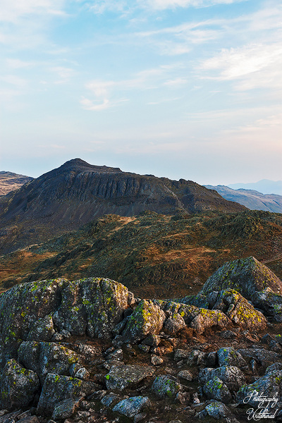 Bowfell links from Crinkle Crags. Rugged mountain landscape with mossy rocks in the foreground, under a blue sky with wispy clouds. The scene conveys a serene, natural beauty.