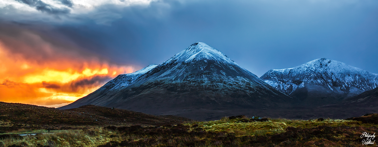 Snow-capped mountains under a dramatic sky, with a fiery sunset on the left and dark clouds above. Foreground features a grassy landscape. Majestic and moody. Glamaig: Isle of Skye. Landscape Photography on the cheap.