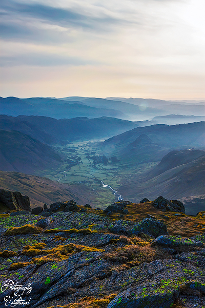 Langdale Valley. A vast mountain landscape at sunrise, with sunlit valleys and a winding river. The foreground features rocky terrain with patches of green moss.