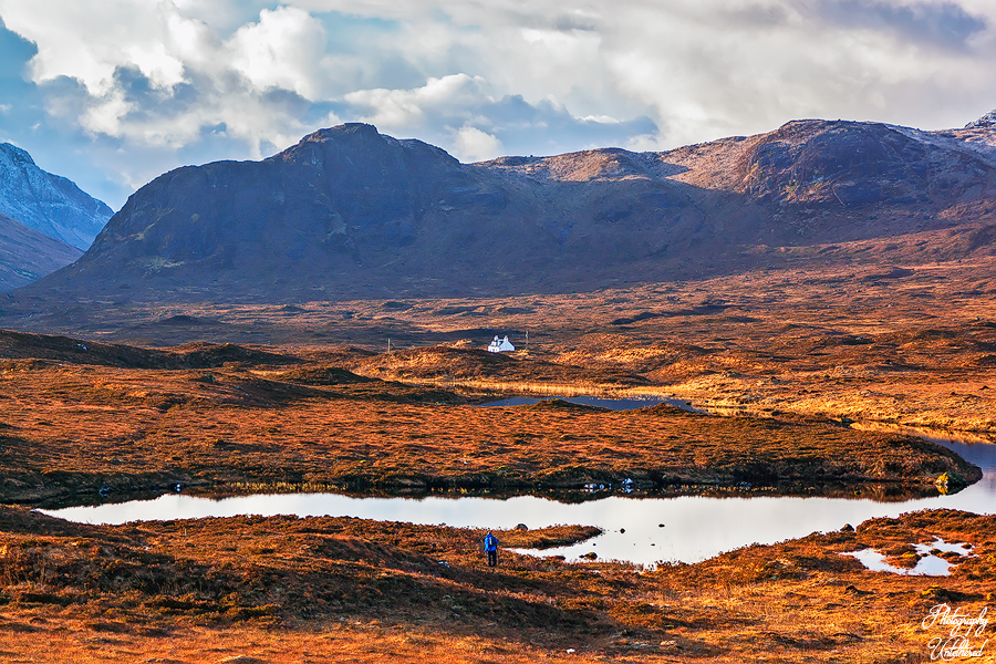 A person walking along a path with a mountain backdrop, surrounded by greenery and clear blue skies. Isle of Skye