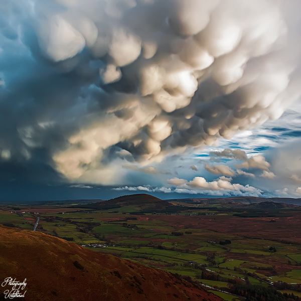 Dramatic, bulbous clouds loom over a landscape of green fields and rolling hills, creating a serene yet ominous atmosphere under the expansive sky. Mammatus Clouds from Scales Fell. Lake District Photography.
