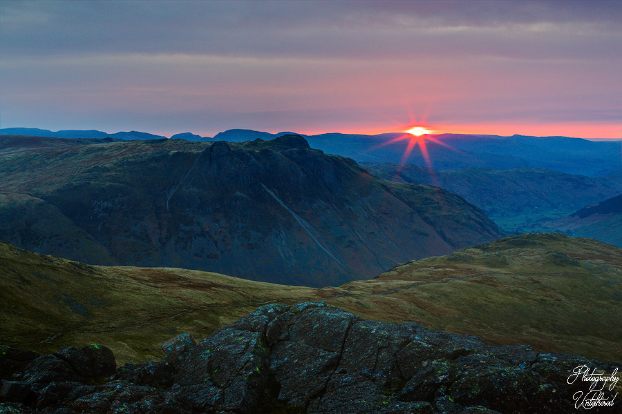 Sunrise over rolling hills and rocky terrain, with a vibrant red sun rising behind distant mountains under a cloudy sky, creating a serene, majestic scene. Pike of Stickle.