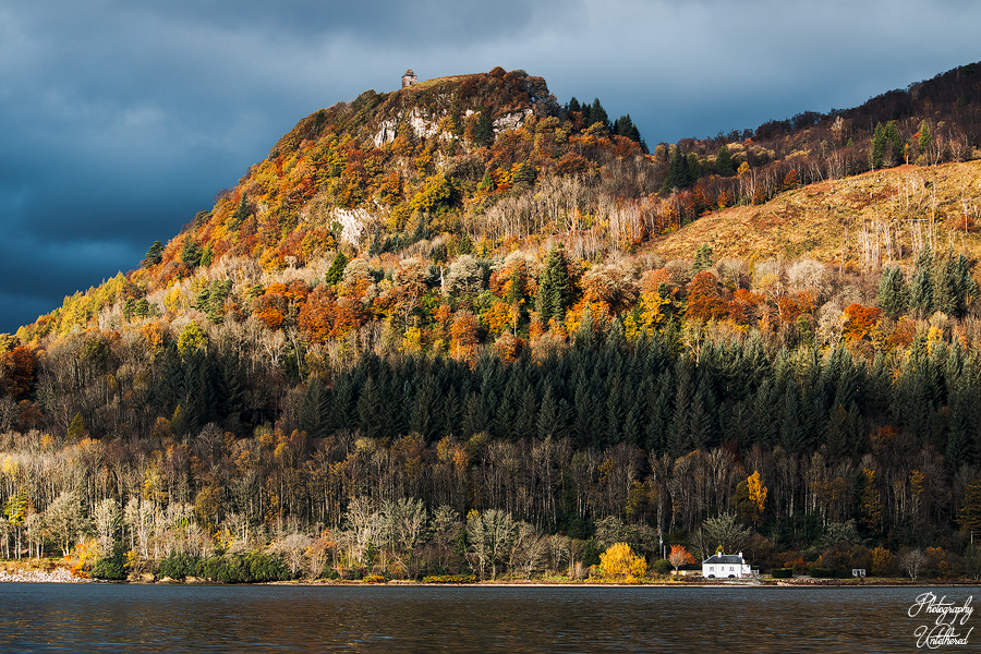 Scenic view of a forested hill in autumn, with vibrant orange and yellow foliage. A small white house sits by a calm lake under a dramatic, cloudy sky. Dùn na Cuaiche, Inveraray. Photography Untethered.