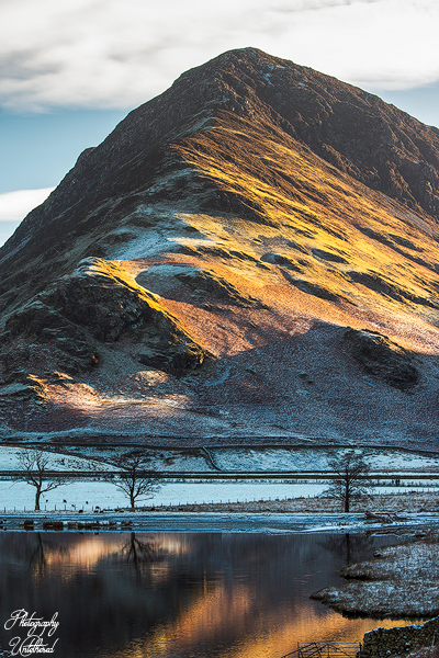 A dramatic mountain with patches of sunlight and shadow towers over a snow-dusted landscape. A still lake reflects the scene, evoking tranquillity and awe. Fleetwith Pike, Lake District. Lake District Photography.