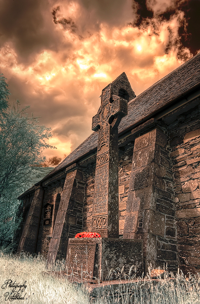 A tall, intricately carved Celtic cross stands before a stone church under dramatic, cloudy skies. The scene evokes a somber, historical atmosphere. Holy Trinity Church Memorial Cross Borrowdale Lake District