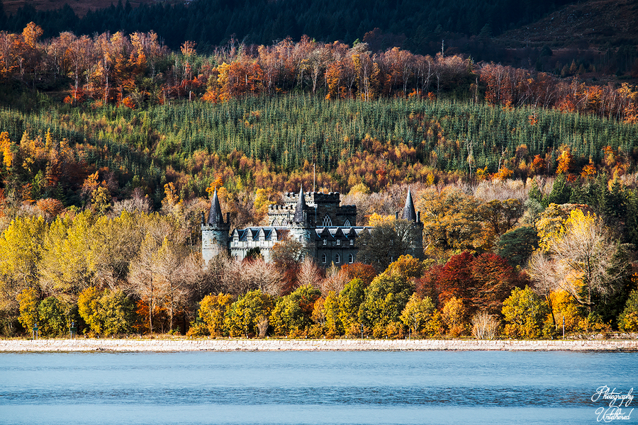 Inveraray castle with tall spires stands amidst a forest with vibrant autumn foliage in shades of orange, yellow, and red, next to a calm blue lake. Landscape Photography Scotland.