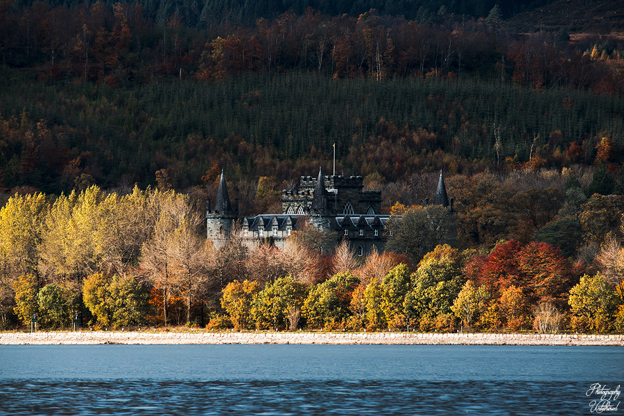 A distant Inveraray Castle with turrets is nestled in a dense forest of colorful autumn trees, beside a serene blue lake, evoking a mystical, tranquil atmosphere. Landscape Photography Scotland.