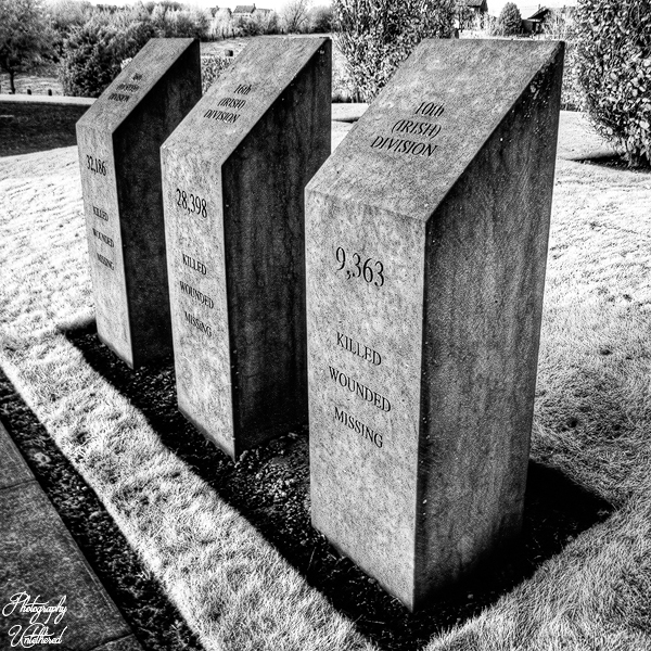 Three stone memorial pillars stand in a line on a grassy field, each inscribed with division names and casualty numbers, evoking somber reflection. Irish Peace Park.
