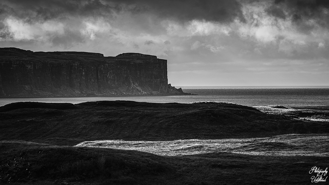 Black and white coastal landscape with dramatic cliffs and ocean under a cloudy sky. Rolling hills in the foreground add depth and texture. Minginish Peninsula and Loch Bracadale Isle of Skye. Black and White Photography.