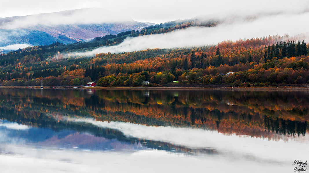 A serene lake reflects autumn-colored trees and misty mountains in a tranquil landscape. The scene conveys peace and natural beauty, with a soft, gentle tone. Loch Eil. Photography Untethered.