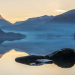 Serene Ullswater lake at sunset, with misty blue mountains and a golden sky. Large rocks in the foreground reflect the calm water, evoking tranquillity. Lake District Photography.