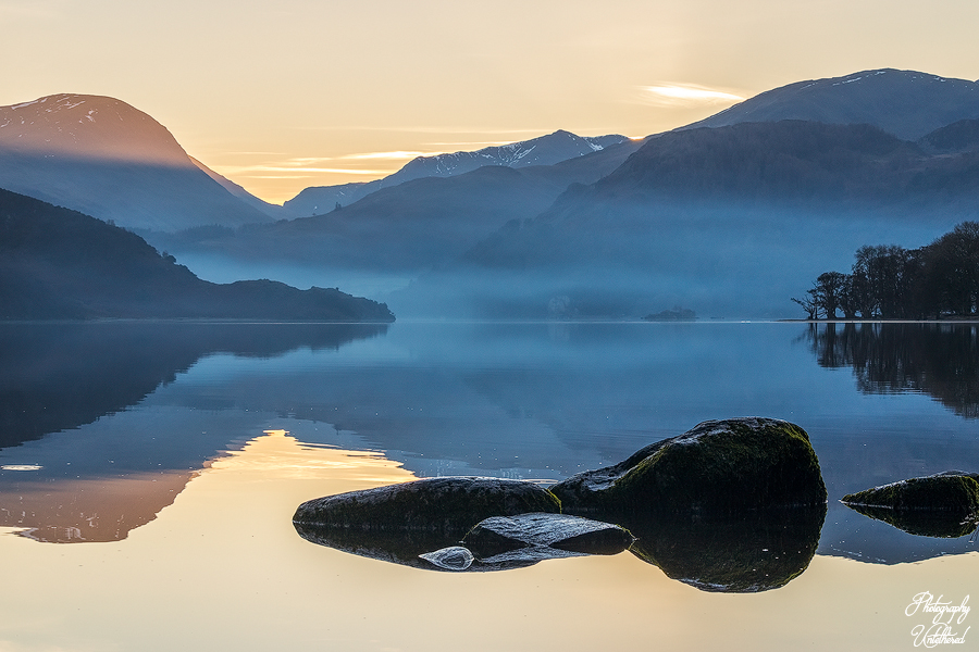 Serene Ullswater lake at sunset, with misty blue mountains and a golden sky. Large rocks in the foreground reflect the calm water, evoking tranquillity. Lake District Photography.