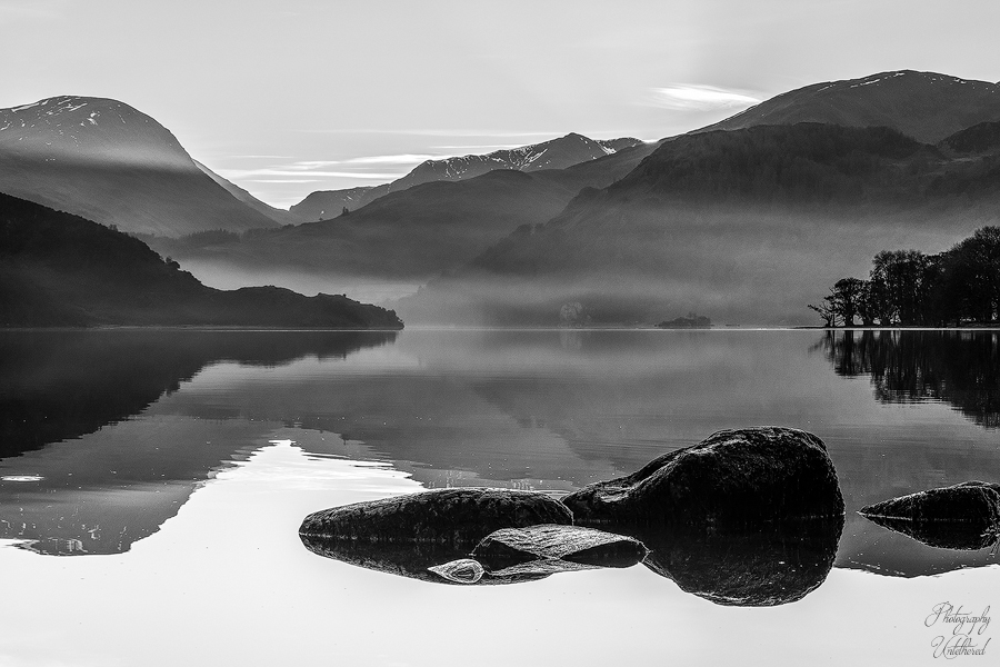 A serene black-and-white Ullswater landscape shows a misty lake reflecting mountains and the sky. Foreground rocks add texture, creating a calm, tranquil atmosphere. Black and White Photography.
