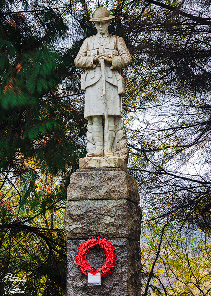 A stone soldier statue stands on a pedestal surrounded by trees. A red wreath is attached to the base. The scene conveys a solemn, respectful tone. Glenfinnan WW1 War Memorial