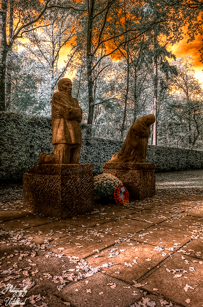 Two sombre stone statues stand amid fallen leaves, with one figure upright and another bowed. The sky glows orange, creating a surreal atmosphere. The Grieving Parents Vladslo German War Cemetery. Käthe Kollwitz.
