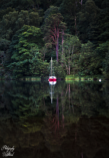A red sailboat is anchored on a still, reflective lake surrounded by dense green forest. The peaceful scene evokes tranquillity and solitude.St Patrick's Boat Landing Ullswater. Lake District Photography.