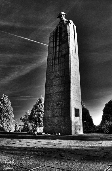 Tall stone monument with a soldier sculpture at the top against a dramatic sky. The word "Canada" is engraved, surrounded by trees, evoking solemnity. St. Julien Memorial, also known as The Brooding Soldier.