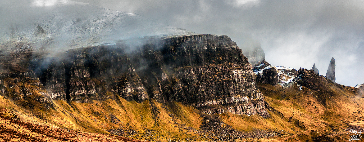 Rugged mountain landscape with dramatic cliffs shrouded in mist, contrasting the golden-brown grassy slopes below. Moody, atmospheric scene. An Stòr Skye.